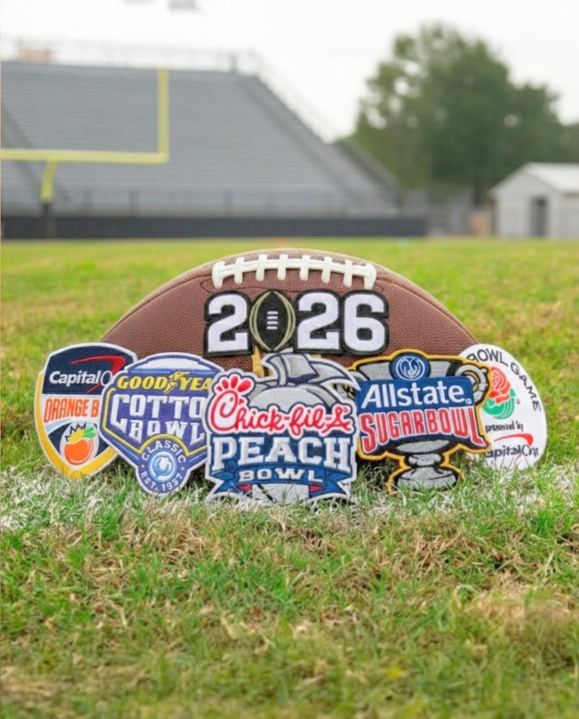 Football-shaped sign with 2026 Super Bowl logo and various bowl game logos on a grassy field.