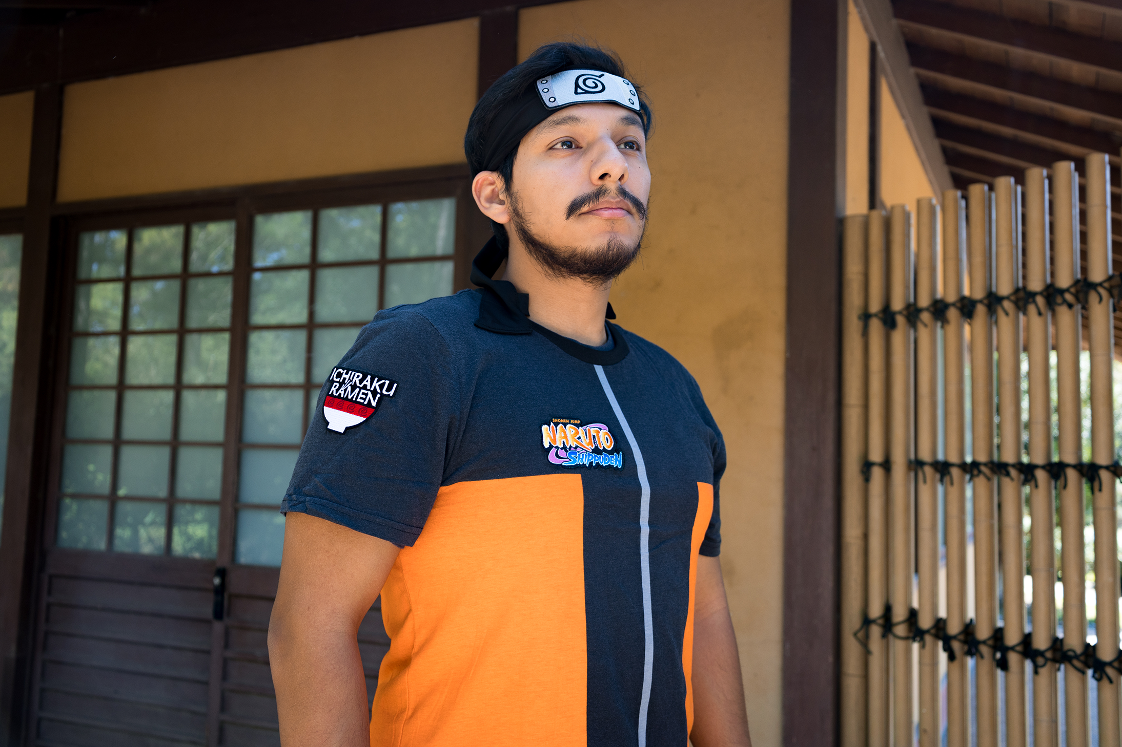 Person wearing a black and orange shirt with logos, standing in front of a traditional wooden building.