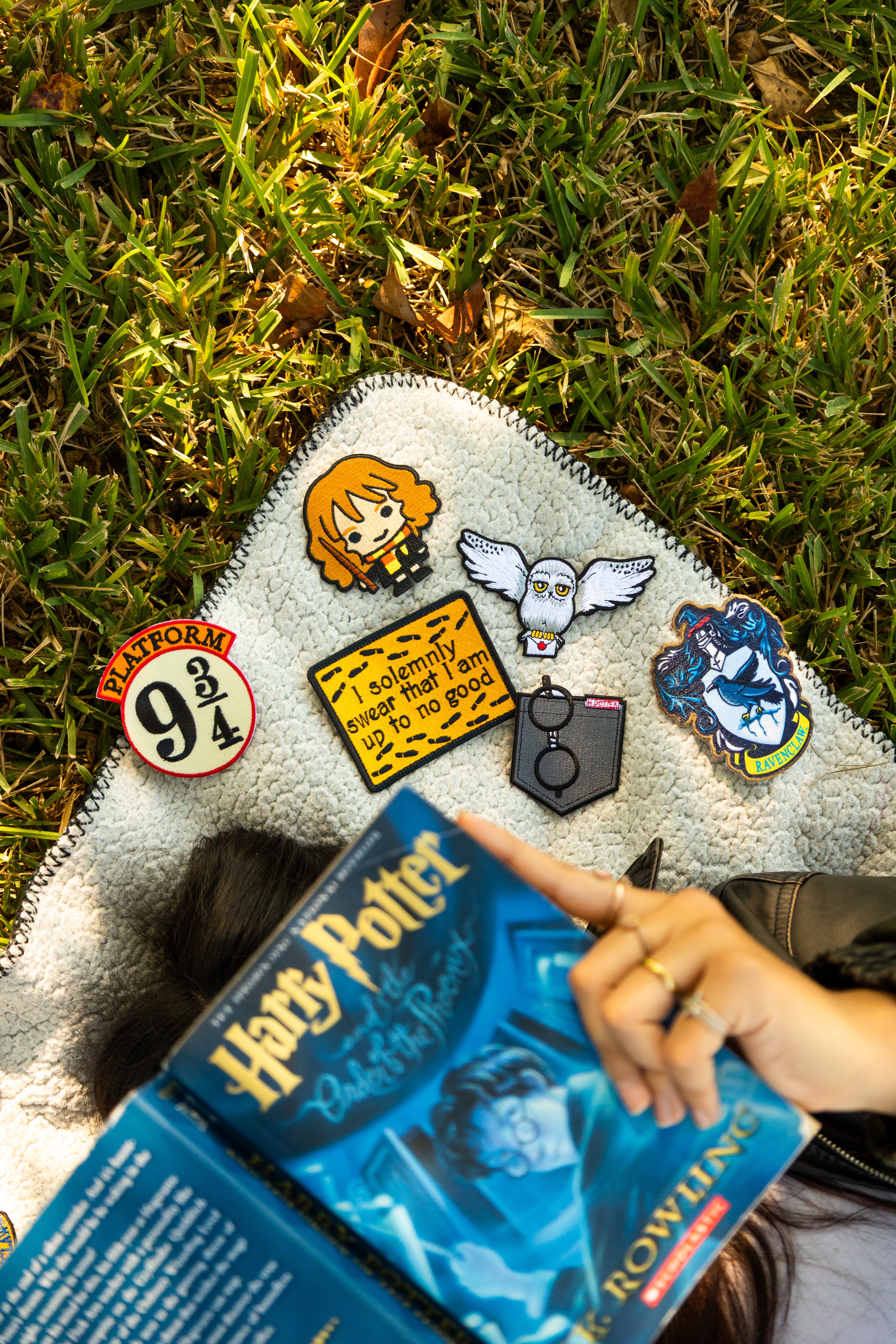 Person holding a 'Harry Potter' book with a jacket featuring character pins on grass
